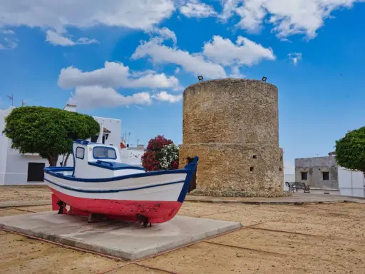 Ein Fischerboot und ein alter Turm in der conil altstadt. Weiße Häuser und grüne Bäume unter blauem Himmel mit Wolken.
