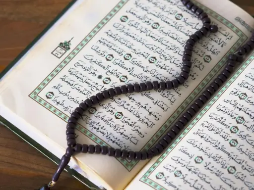 Ein Koran mit Perlenkette liegt auf einem Holztisch. Die Seiten sind mit arabischer Schrift bedruckt, die an die Geschichte von Yajuj und Majuj im Islam erinnert.