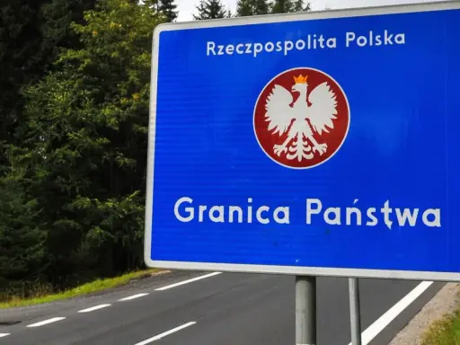 Niebieski znak z orłem i napisem "Rzeczpospolita Polska" oraz "Granica Państwa" stoi przy drodze otoczonej lasem.