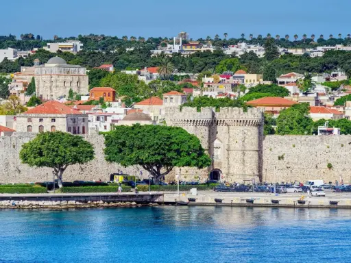 Widok na zabytkowe mury obronne i zabudowania wyspy Rodos, z błękitnym morzem w tle.