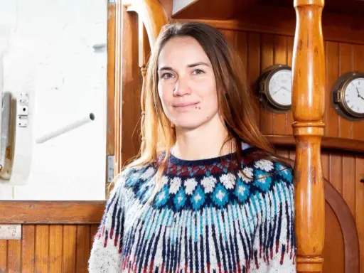 Laura Dekker heute, lächelnd in einem bunten Strickpullover, an Bord eines Schiffes mit Holzakzenten und maritimen Uhren.