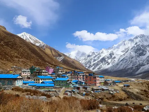Villaggio colorato ai piedi dell'alta montagna himalayana, con case dai tetti blu e rosa, circondato da pendii bruni e cime innevate.