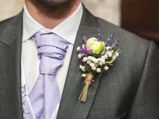 Elegancka butonierka w marynarce, ozdobiona białymi kwiatkami i lawendą, dodaje uroku ślubnemu strojowi.