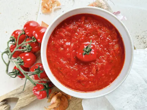 Eine Schüssel mit köstlicher, hausgemachter italienischer Tomatensauce, garniert mit einer frischen Kirschtomate. Daneben liegen frische Tomaten und Zwiebeln.