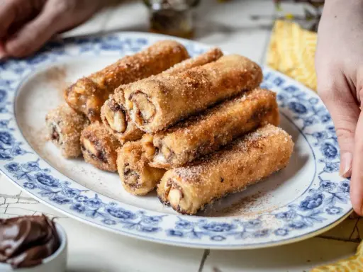 Nutella French Toast Rolls, goldbraun gebacken und mit Zimt-Zucker bestreut, auf einem Teller mit blauem Blumenmuster.