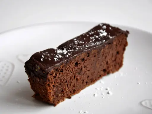 Wilgotny brownie z czerwonej fasoli, polany czekoladą i posypany cukrem pudrem, na białym talerzu z serduszkami.