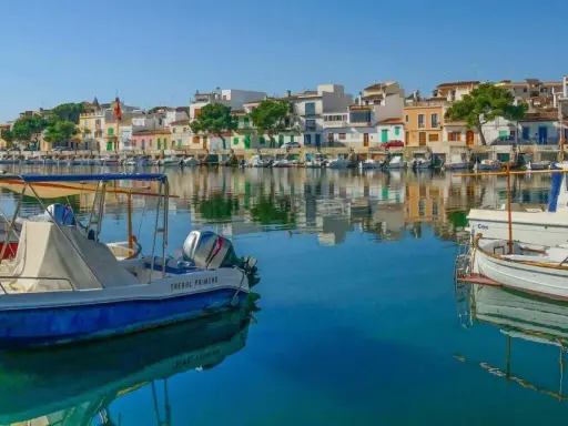 Boote im Hafen von Portocolom, Mallorca, spiegeln sich im ruhigen, blauen Wasser vor bunten Häusern.