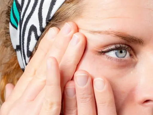 Doigts appliquant un soin pour le massage cernes, près d'un œil bleu. Bandeau à rayures zébrées.