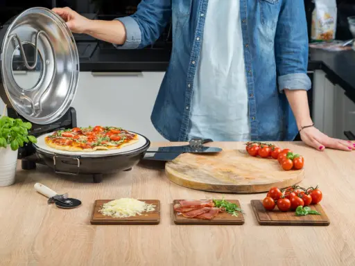 Otwarta opiekacz do pizzy, na kamieniu piec pizza z pomidorkami i bazylią. Obok deska z pomidorkami, startym serem i szynką.