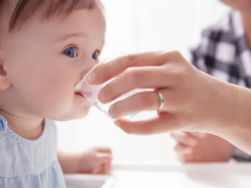Ein Baby trinkt vorsichtig Wasser aus einem Becher, gehalten von einer Hand. Die Frage "ab wann Baby Wasser geben?" wird hier visuell beantwortet.