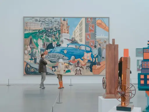 Two people observe a large, colorful mural depicting a chaotic scene with a blue car, factories, and people, offering a glimpse into the art market definition of diverse artistic expression.