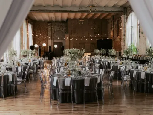 Eleganckie wesele w industrialnym stylu. Stoły nakryte szarymi obrusami, ozdobione białymi kwiatami i transparentnymi krzesłami. Girlandy świateł tworzą magiczny nastrój.