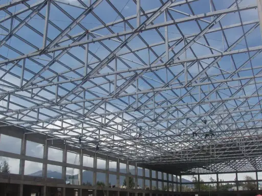 Estructura metálica de una **cercha Pratt** en construcción, con operarios trabajando en el techo bajo un cielo azul.