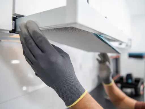 Hände mit Handschuhen ziehen einen Filter aus einer Dunstabzugshaube, um den Motor zu reinigen.