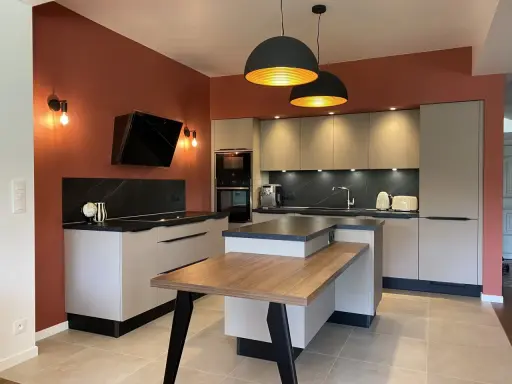 Cuisine moderne aux murs terracotta, avec îlot central en bois et mobilier gris clair. Éclairage chaleureux.