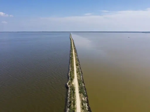 Un sentiero stretto divide due specchi d'acqua, uno scuro e uno più chiaro, sotto un cielo azzurro. Sembra la ciclabile degli angeli.