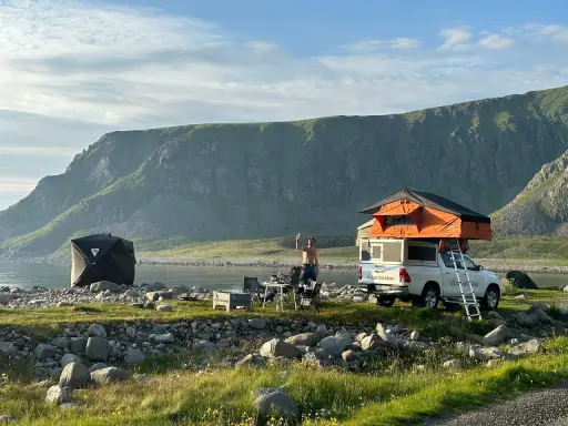 En man tar en selfie vid sitt tak-tält på en strand i Norge. Bredvid står en mörk kupoltält och ett bord med stolar. Berg i bakgrunden.