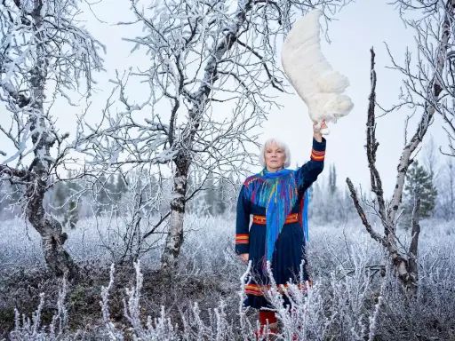 Britta Marakatt Labba i samedräkt håller upp en stor vit fjäder i en snötäckt skog.