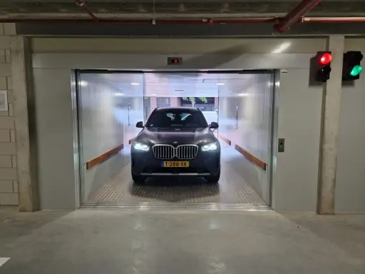 Un coche BMW oscuro espera en un montacoches para garaje. La luz roja del semáforo indica que debe esperar.