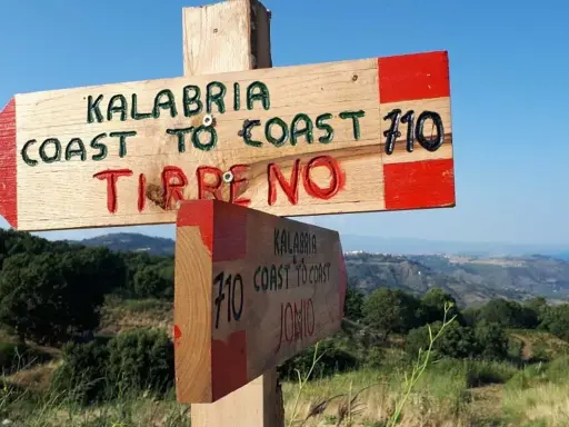 Cartello in legno indica "Kalabria Coast to Coast" verso il Tirreno e lo Jonio, con sfondo di colline e mare.