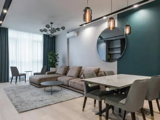 Nowoczesna lampa do salonu, wisząca nad stołem, tworzy przytulną atmosferę. W tle duża sofa i okno z zasłonami.
