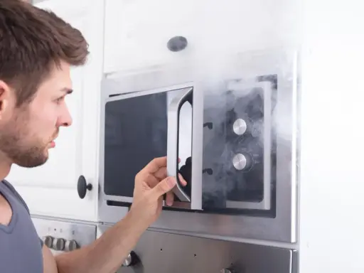 Mann öffnet Mikrowelle, aus der Rauch dringt. Ein Fehler mit Metall in Mikrowelle kann gefährlich sein.