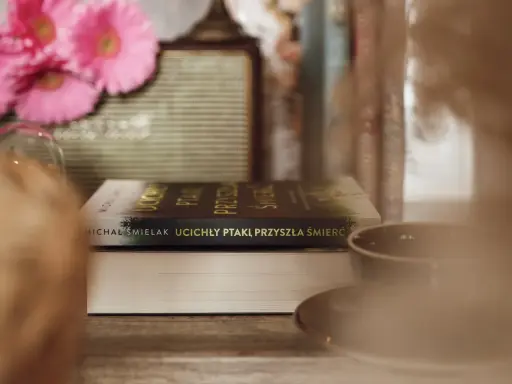 Na stole leżą książki, jedna z nich ma tytuł "Ucichły ptaki, przyszła śmierć". W tle różowe kwiaty i filiżanka.