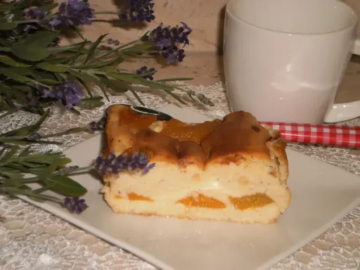 Pyszny sernik z brzoskwiniami, udekorowany gałązką lawendy, obok białej filiżanki i czerwono-białej łyżeczki.