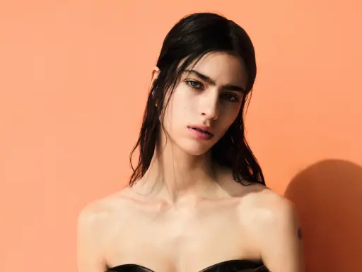 A striking portrait of Loli Bahia, her dark hair cascading around her shoulders against a vibrant peach backdrop.