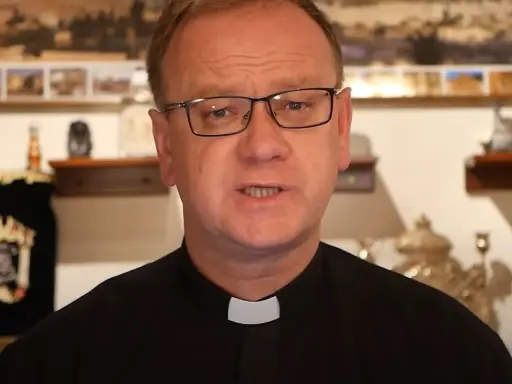 Wojciech Węgrzyniak w okularach, w czarnej sutannie z białą kolaretką, przemawia. W tle widać zwoje Tory i obrazy.
