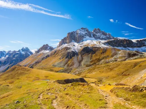 Ampia **foto grandangolo** di montagne innevate e pendii dorati autunnali, con un sentiero che serpeggia tra l'erba.