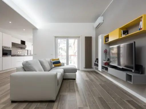 Un progetto soggiorno stretto e lungo con divano grigio, TV a parete e mensole gialle. La cucina moderna è visibile sullo sfondo.