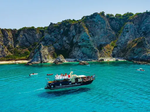 Barca turistica naviga nelle acque cristalline vicino al porto di Tropea, con imponenti scogliere a picco sul mare e piccole spiagge affollate.