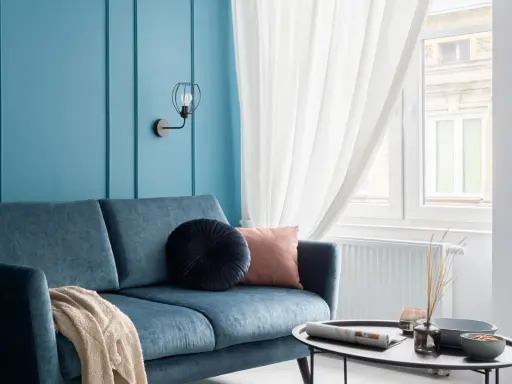Divano blu petrolio con cuscini decorativi e coperta. Parete azzurra con applique. Abbinamenti moderni e rilassanti.