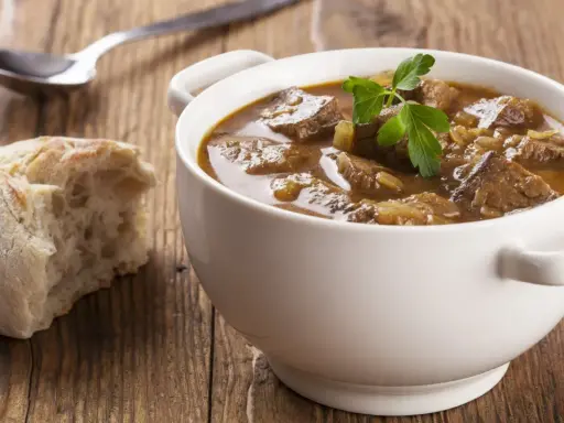 Aromaticzny gulasz wołowy w białej miseczce, ozdobiony natką pietruszki, obok kromka chleba i łyżka.