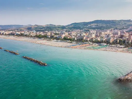 Vista aerea di San Benedetto del Tronto: una lunga spiaggia con ombrelloni colorati e il mare turchese, con dighe frangiflutti che proteggono la costa.