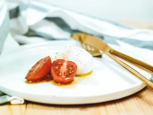 Kuleczki mozzarelli z pomidorkami koktajlowymi na białym talerzu, obok złote sztućce i ściereczka.