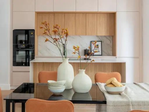 Dos jarrones blancos con flores anaranjadas y amarillas, cuencos apilados y un bol con panecillos son elementos decorativos que poner encima de una mesa de comedor.