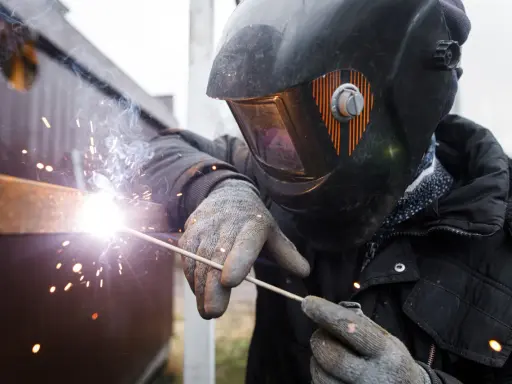 Soldador con casco y guantes usa un electrodo para unir metales, creando chispas brillantes y humo.
