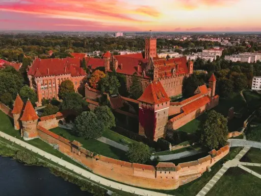 Zamek krzyżacki w Malborku, imponująca twierdza z czerwonej cegły, otoczona fosą i zielenią, pod rozgwieżdżonym, różowym niebem. Ciekawostki o jego historii czekają na odkrycie.