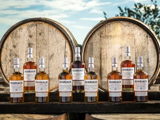 Butelki whisky BenRiach stoją przed beczkami, które zdradzają, skąd pochodzi whisky.