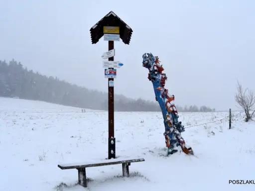 Zimowy krajobraz z ławką i drogowskazem turystycznym. Obok kolorowa rzeźba, być może symbolizująca skopiec szczyt.
