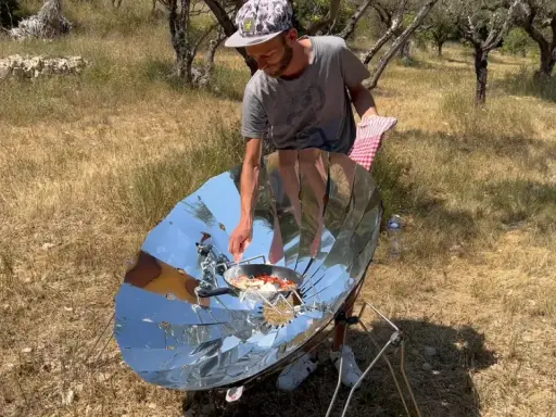Un homme cuisine avec un four solaire parabolique, démontrant le fonctionnement de cet appareil écologique en plein air.