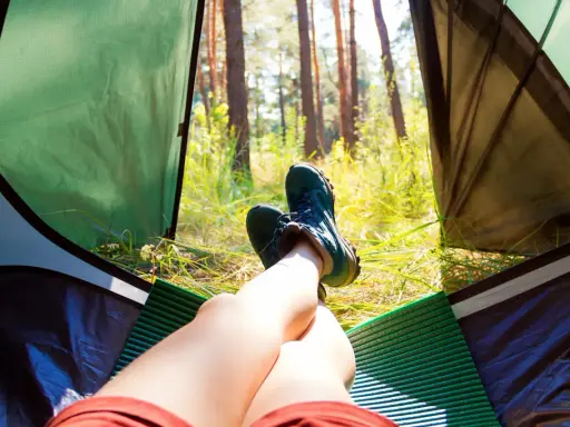 Nogi w butach trekkingowych odpoczywają w namiocie. Słońce przebija się przez drzewa. Idealne miejsce, by zanocuj w lesie.