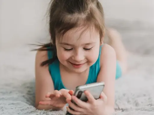 Uśmiechnięta dziewczynka leży na dywanie, bawiąc się telefonem. Wpływ telefonu komórkowego na mózg dziecka jest tematem dyskusji.