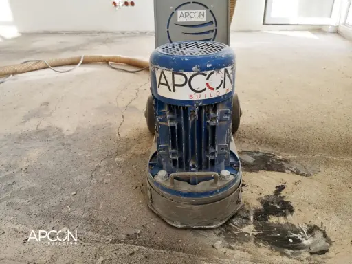 Maszyna APCON przygotowuje podłoże pod parkiet, szlifując beton. Wąż odkurzacza leży obok.