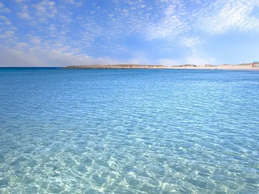 Crystal clear turquoise water with gentle ripples, leading to a sandy beach and distant dunes under a blue sky with scattered clouds.