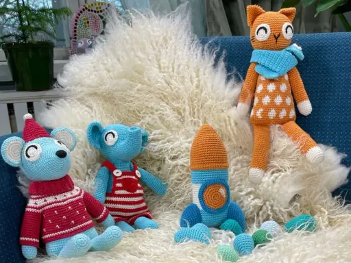 En virkad amigurumi katt med orange och vita prickar sitter på en lurvig matta bredvid en blå mus med tomteluva och en blå elefant i hängselbyxor.