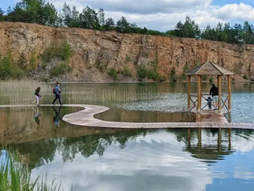 Malownicza trasa Gródka Jaworzno z drewnianym molo i altanką. Dwie osoby spacerują po falistej kładce nad wodą.