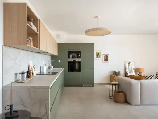 Cuisine moderne avec des armoires vert olive, un plan de travail en marbre et des étagères en bois clair. Un salon adjacent est visible.
